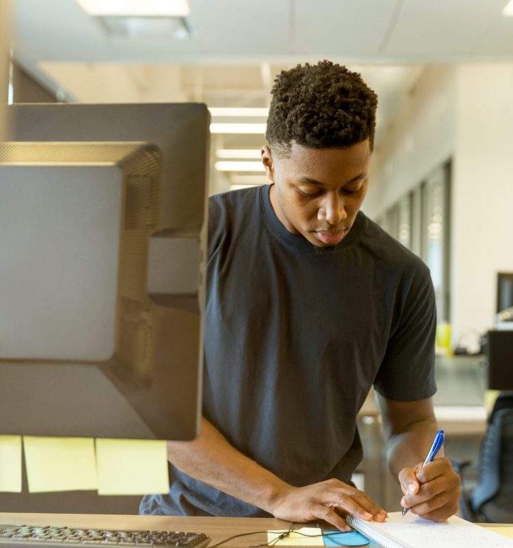 Intern working at a standing desk in front of his computer, writing in a notebook.